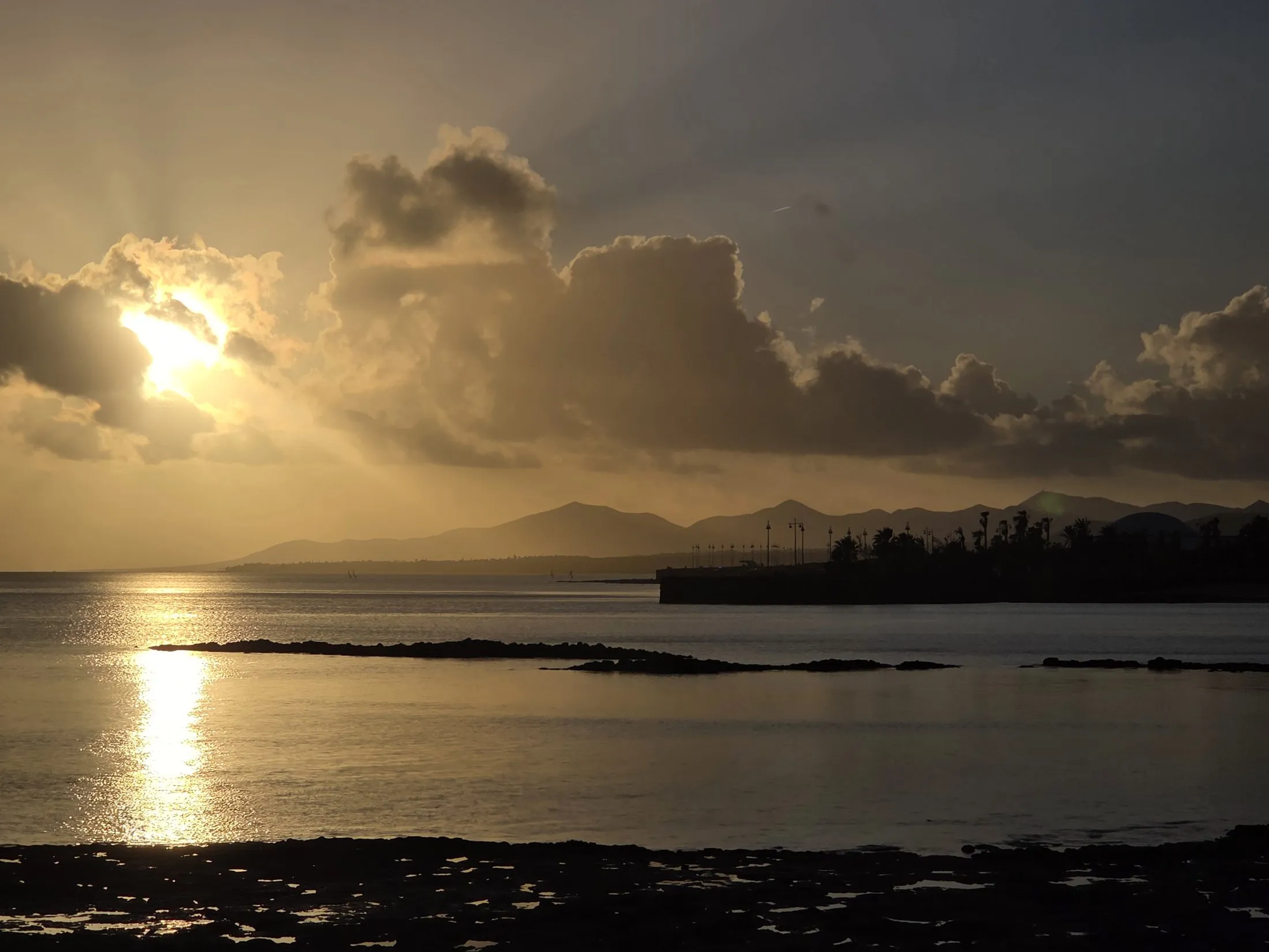 Sun through clouds over a still harbour and distant hills